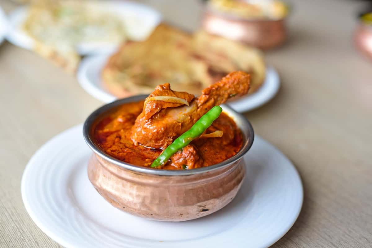 Spicy Indian chicken curry garnished with a green chili, served in a copper bowl with naan bread in the blurred background.