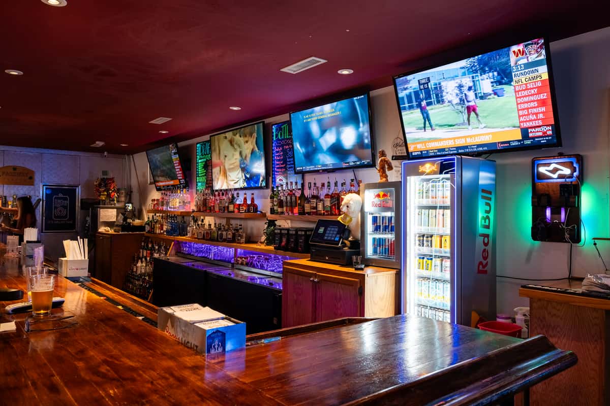 Vibrant bar counter at Parkway Tavern showcasing multiple TVs playing sports, a fully stocked liquor shelf, Red Bull-branded drink coolers, neon lighting, and a polished wooden bar top with beer glasses and cocktail menus.