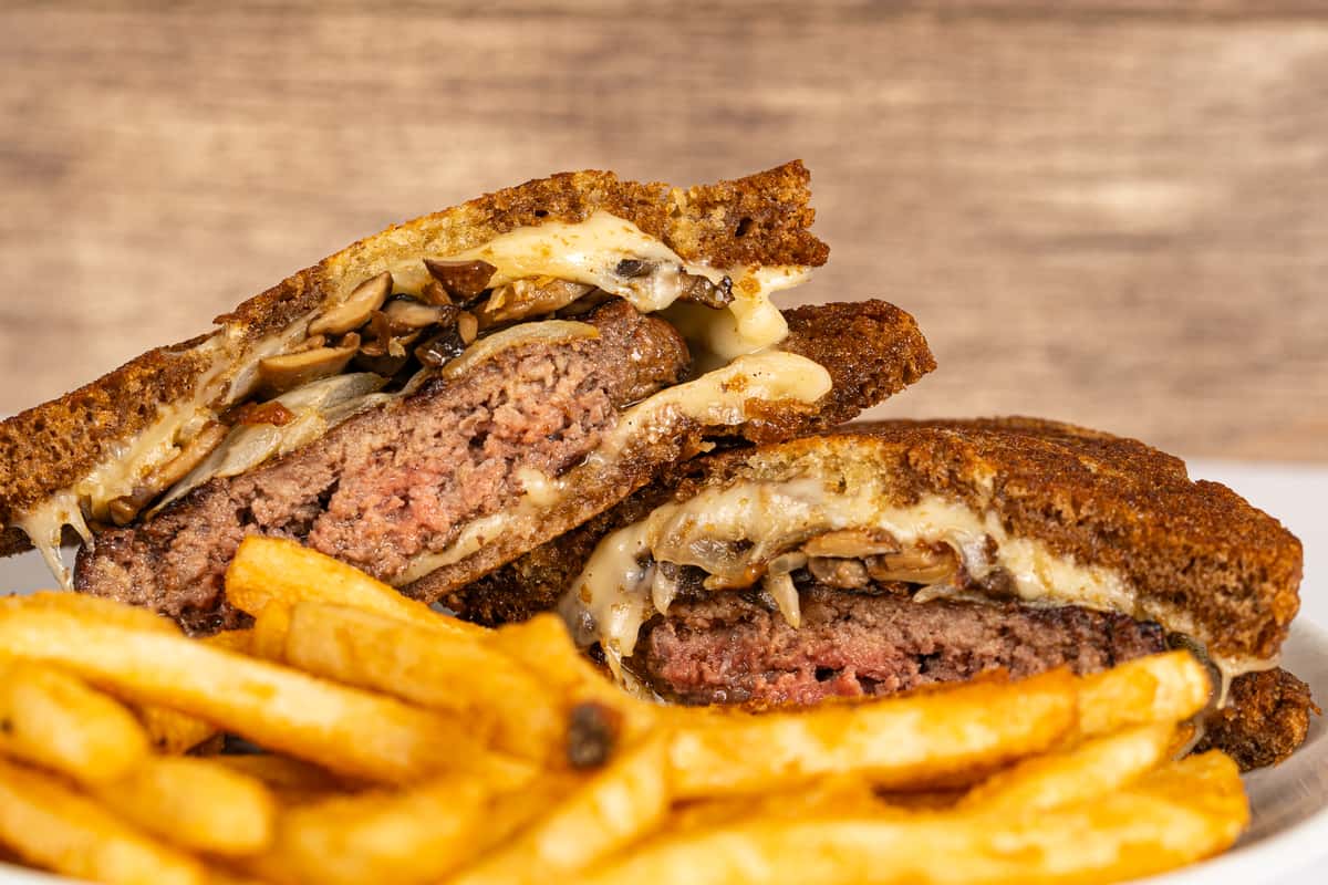 Close-up of a juicy patty melt sandwich stuffed with grilled mushrooms, melted cheese, and beef on crispy toasted rye bread, served with golden French fries on a white plate. Ask ChatGPT