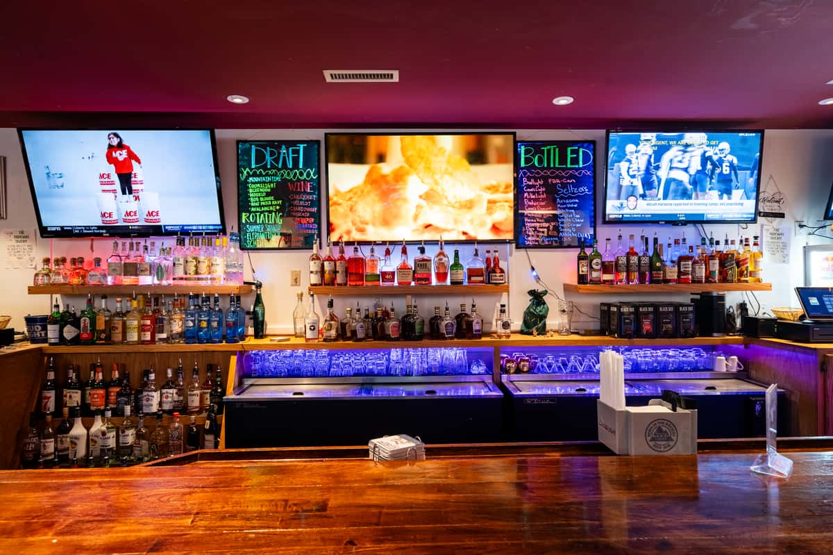 Wide-angle view of the back bar at Parkway Tavern, featuring a colorful liquor display, glowing purple under-shelf lighting, chalkboard menus for draft and bottled drinks, and multiple flat-screen TVs playing sports and food content.