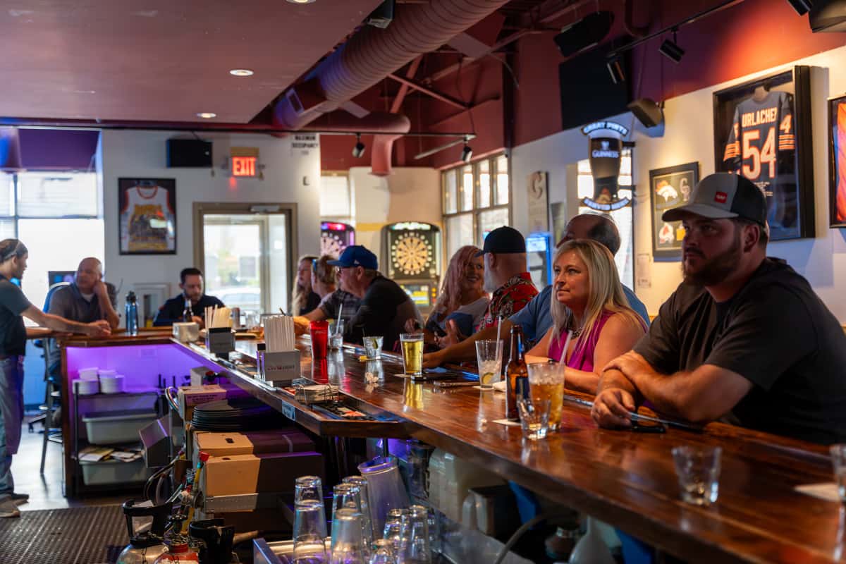 Lively interior of Parkway Tavern with patrons seated along a polished wooden bar, enjoying drinks and conversation in a sports-themed setting with framed jerseys and neon lights.