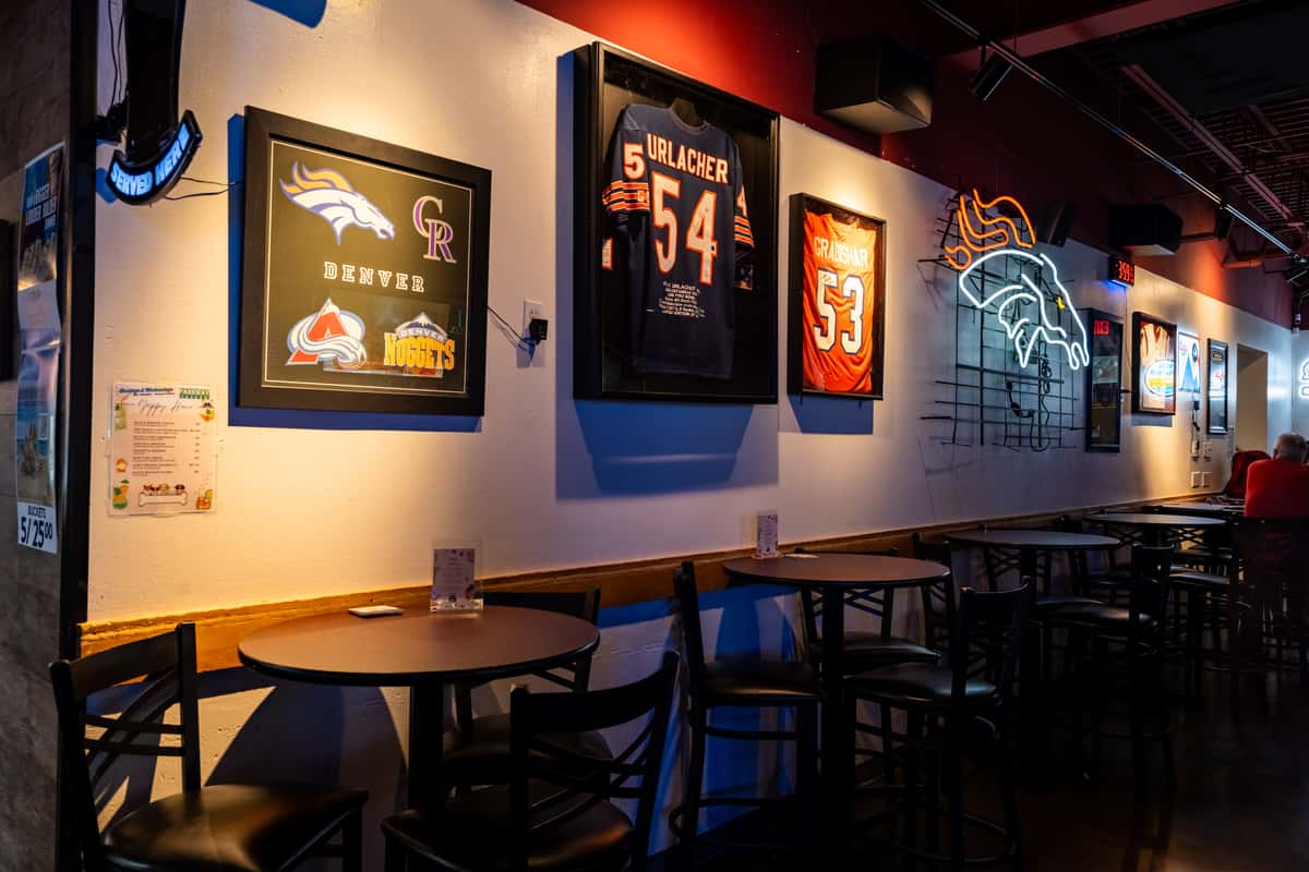 Dimly lit sports bar interior with round high-top tables, framed NFL jerseys, and neon Denver Broncos wall art, highlighting a casual and sporty atmosphere.