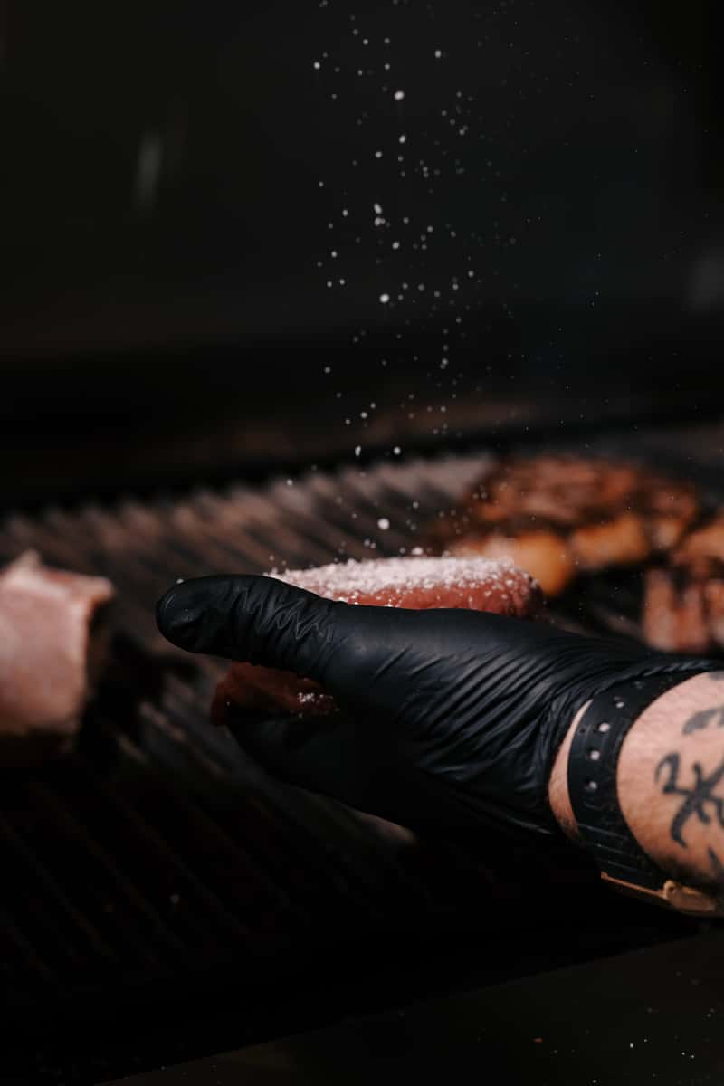 Chef's gloved hand seasoning a raw steak over a grill, salt caught mid-air above the meat.
