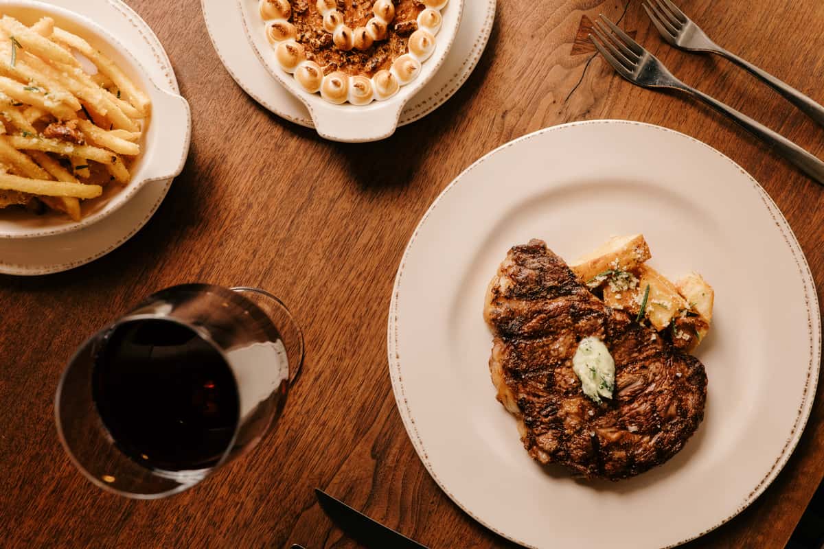 Grilled ribeye steak with herb butter and roasted potatoes, served with truffle fries, sweet potato casserole, and red wine.