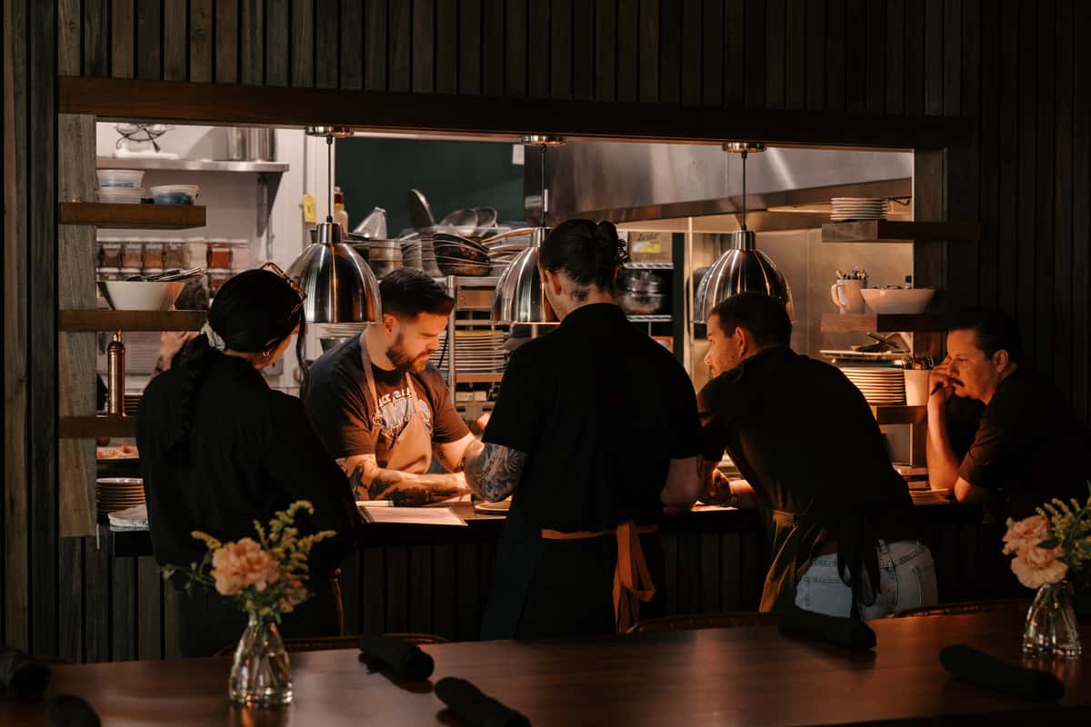 Chefs and kitchen staff gathered at the pass, focused on plating dishes in a warmly lit restaurant kitchen.