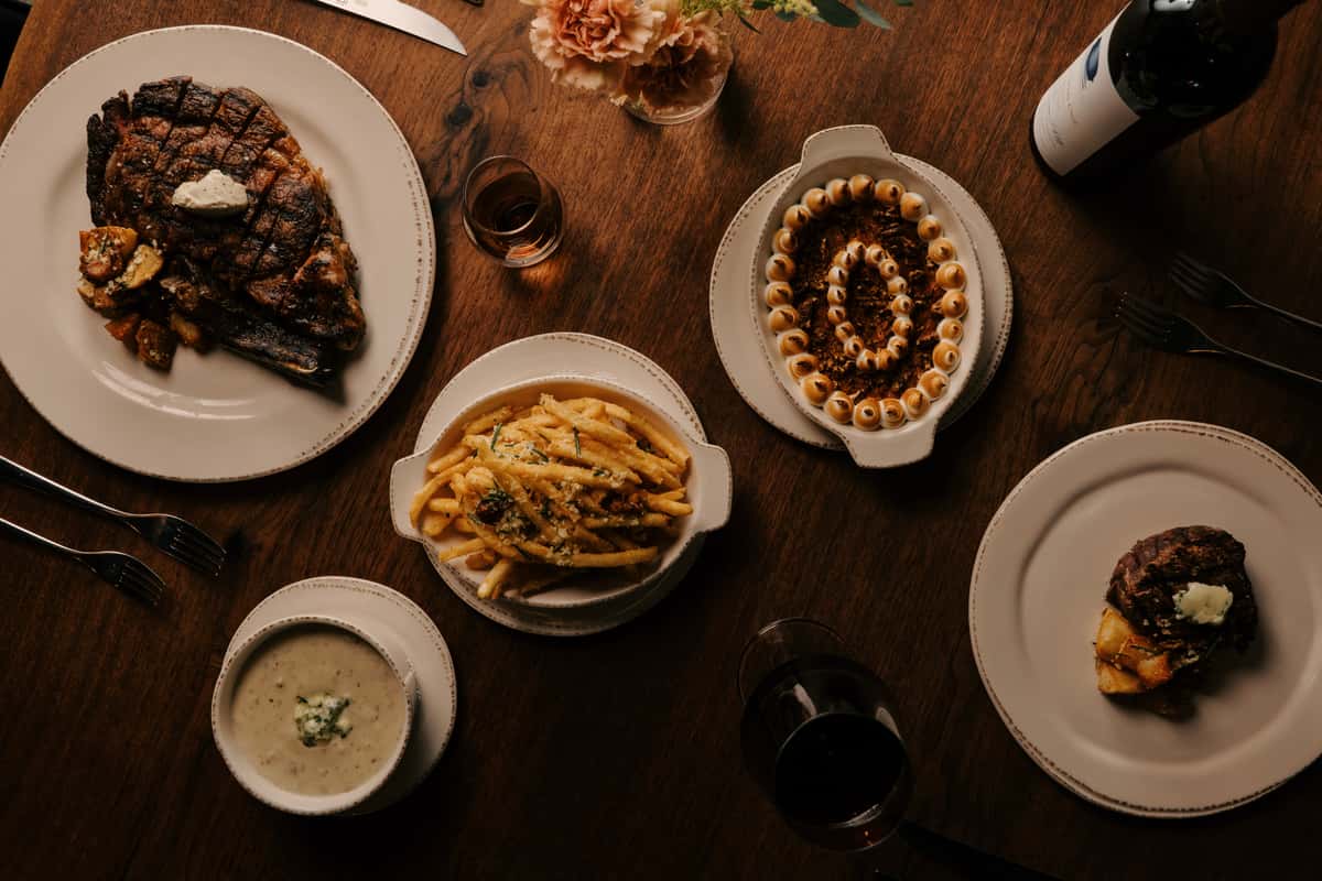 Steakhouse dinner spread with grilled ribeye, filet mignon, truffle fries, soup, sweet potato casserole, and red wine on a wood table.