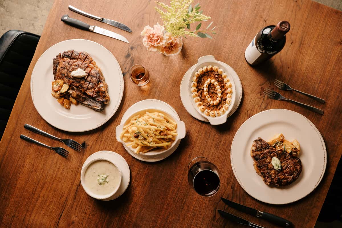 A top-down view of a dinner table featuring a large steak with roasted potatoes, creamy soup, crispy fries, and a dish with a creamy topping, along with a bottle of wine and wine glasses.