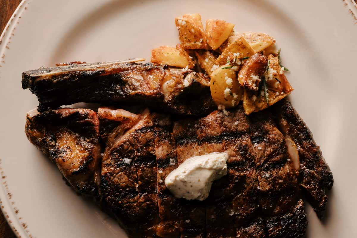 Grilled bone-in steak topped with herb butter, served with roasted potatoes on a white plate.
