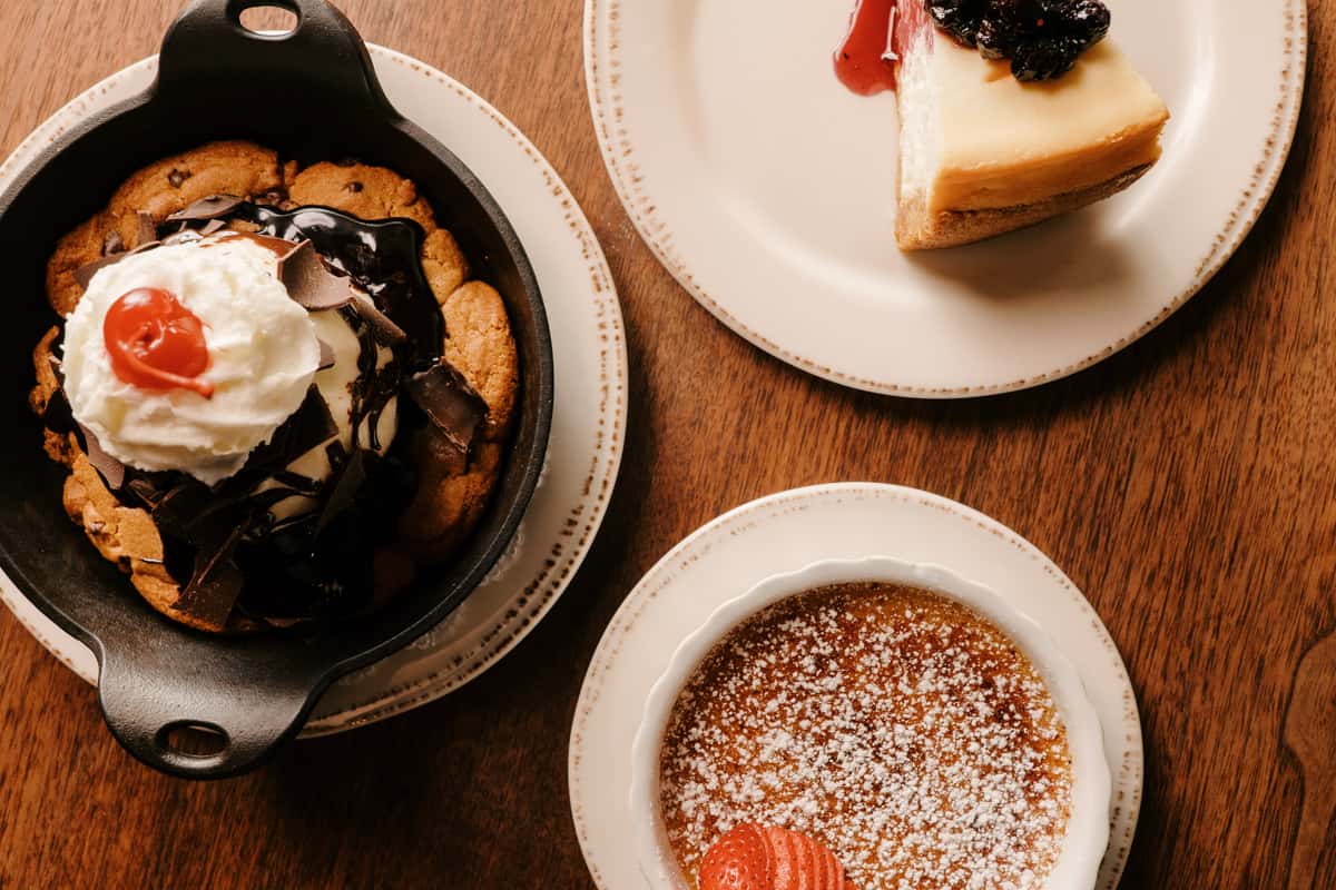 Cheesecake with berry sauce, a chocolate chip cookie sundae, and a chocolate dessert with a strawberry on a wooden table.