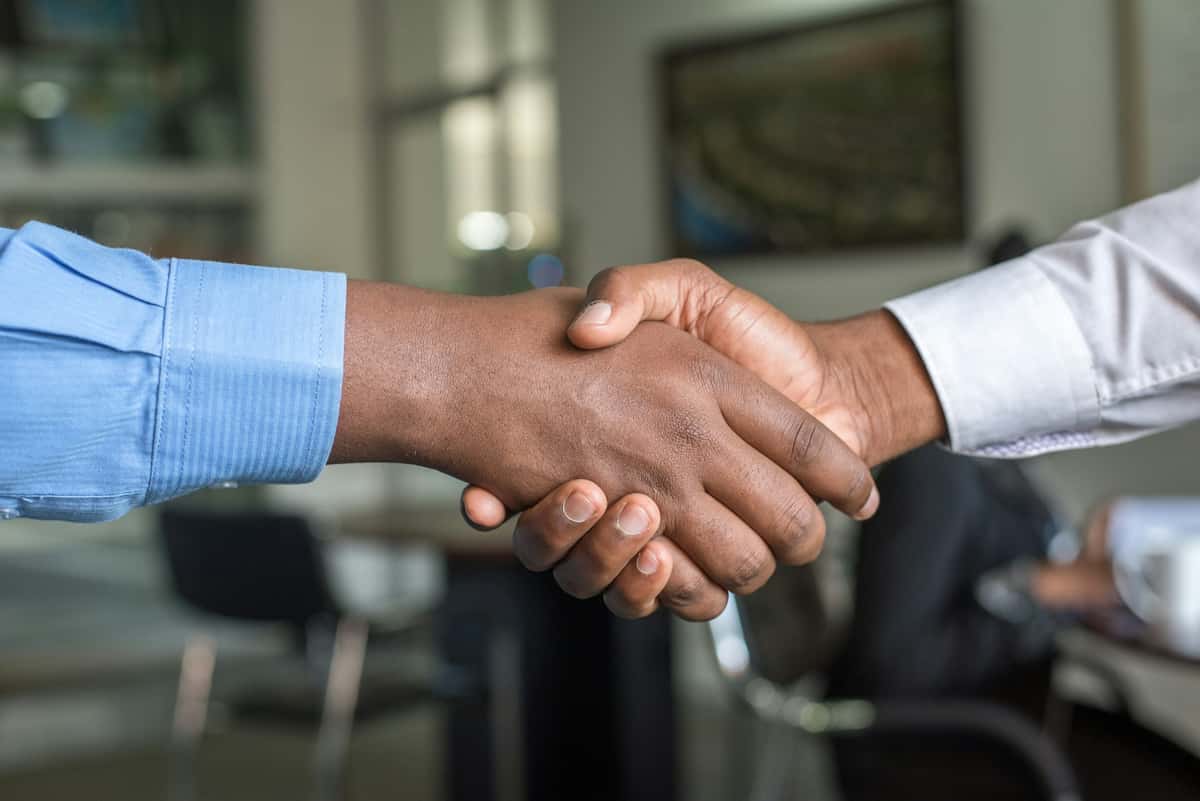 Two dark-skinned hands clasp in a business handshake. The hands are in focus with the blurred background depicting an office setting.