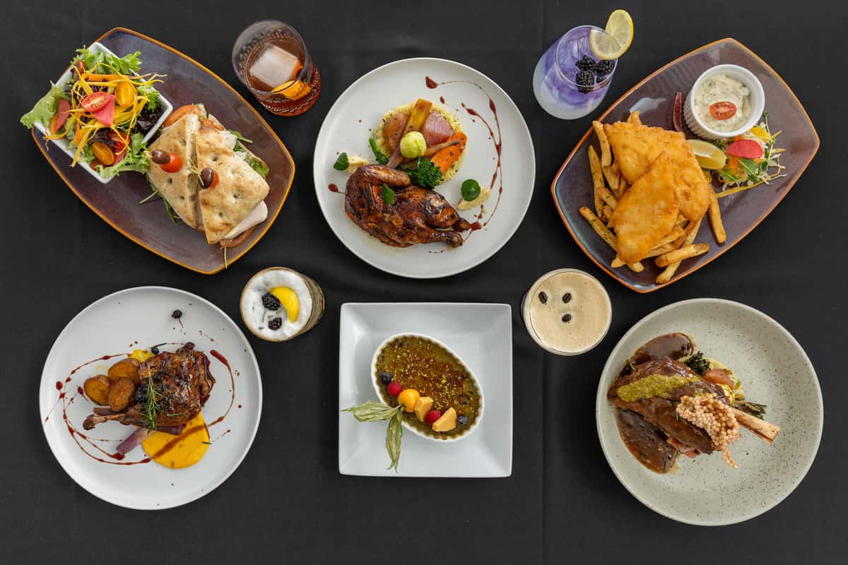 Overhead view of an elegant River View Bistro restaurant table set with gourmet dishes including braised lamb shanks, roasted duck with vegetables, fish and chips with tartar sauce, a fresh focaccia sandwich with mixed greens, and assorted desserts like crème brûlée and a parfait. Accompanied by craft cocktails and coffee-based drinks, all presented on modern white and ceramic plates against a black tablecloth.