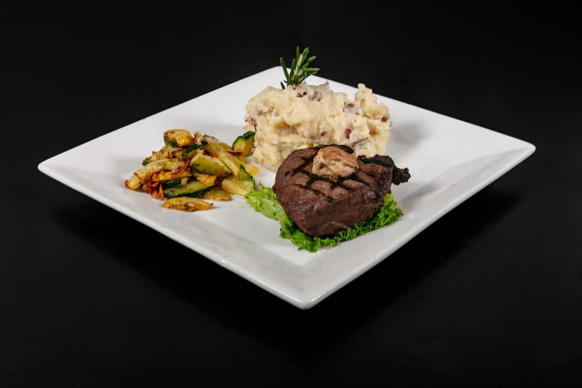 Grilled filet mignon topped with herb butter, served with garlic mashed potatoes and sautéed zucchini and squash on a square white plate.
