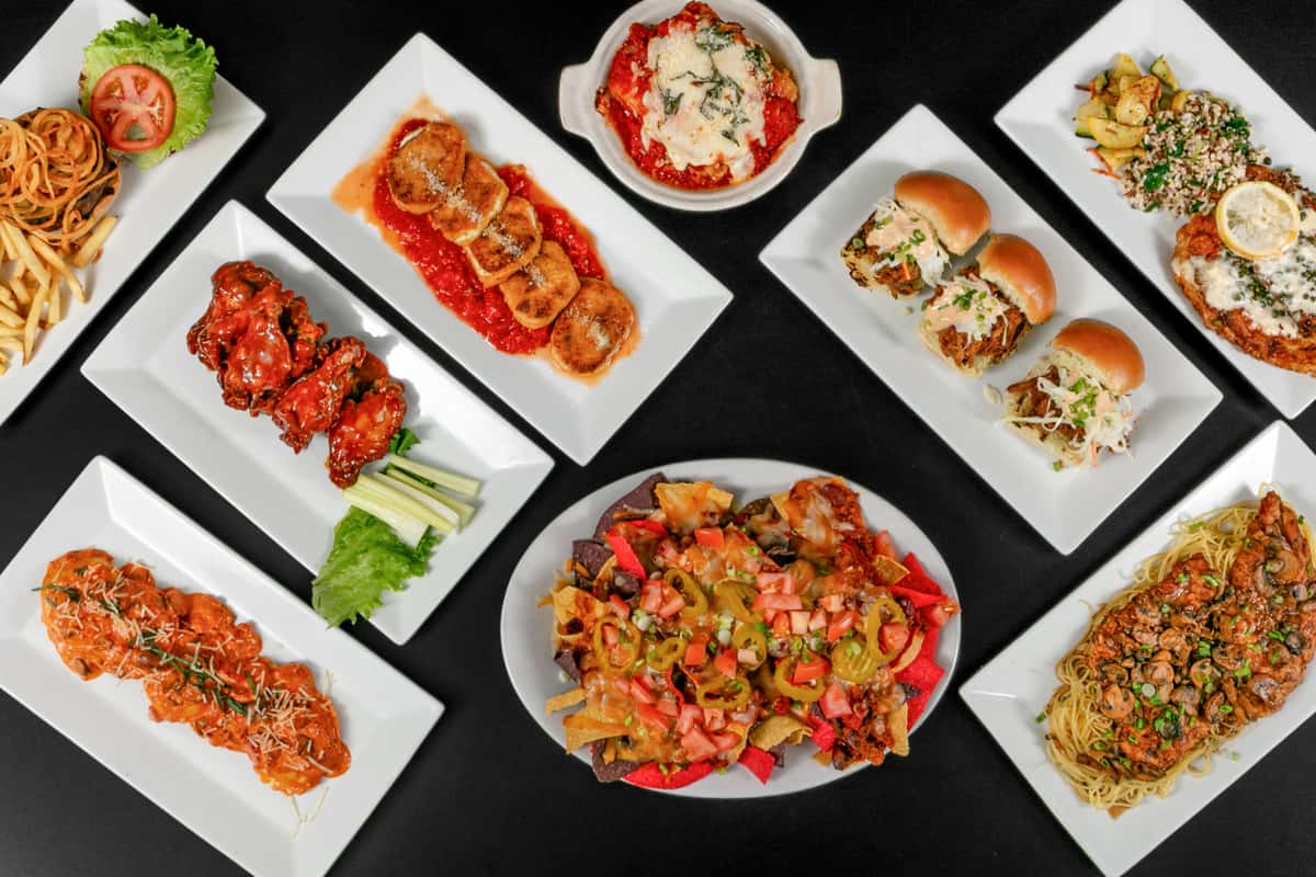 Variety of restaurant dishes on white plates — pasta, sliders, nachos, meat skewers, fried appetizers, and vegetable sides served on a dark table.