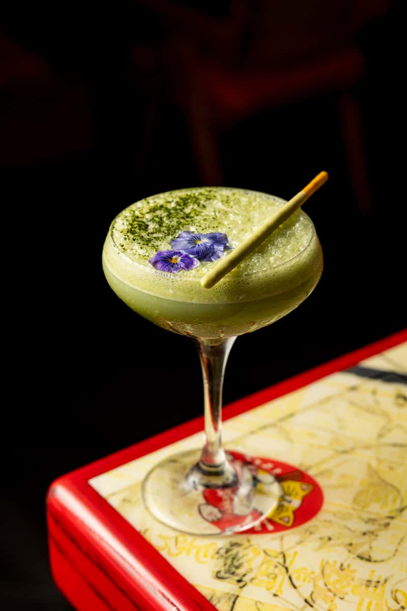 Green tea-inspired craft cocktail garnished with edible flowers at Kapow! Noodle Bar, showcasing creative mixology and elevated Asian fusion dining.
