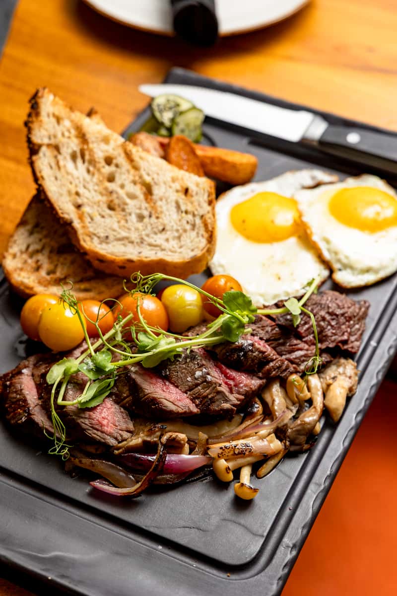 Grilled steak and eggs with roasted tomatoes, mushrooms, and toasted bread at Kapow! Noodle Bar, showcasing a bold modern Asian fusion brunch in Boca Raton and Delray Beach.