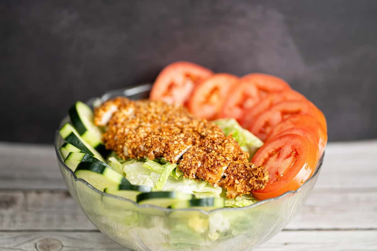 Crispy chicken salad with sliced cucumbers, tomatoes, and shredded lettuce in a clear glass bowl on a wooden surface.