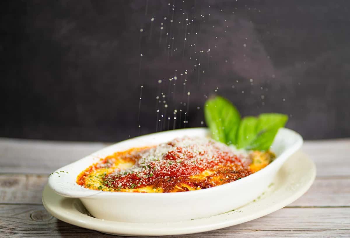 Baked lasagna topped with marinara, grated parmesan, and fresh basil, being sprinkled with cheese on a rustic wooden table.