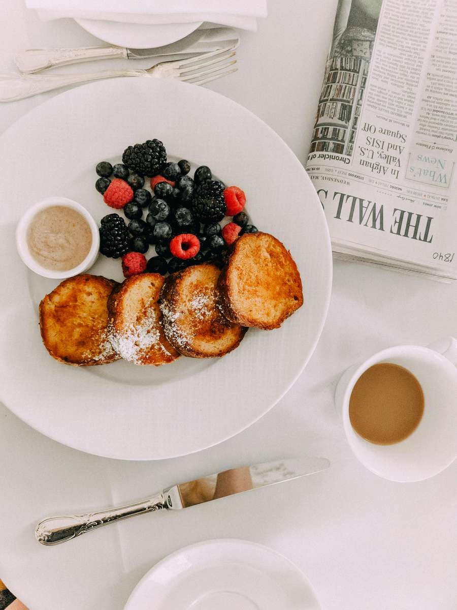 Stylish breakfast setting with golden French toast dusted with powdered sugar, a medley of fresh blackberries, blueberries, and raspberries, and a side of cinnamon dipping sauce, served alongside a cup of coffee and a folded Wall Street Journal on a crisp white tablecloth.