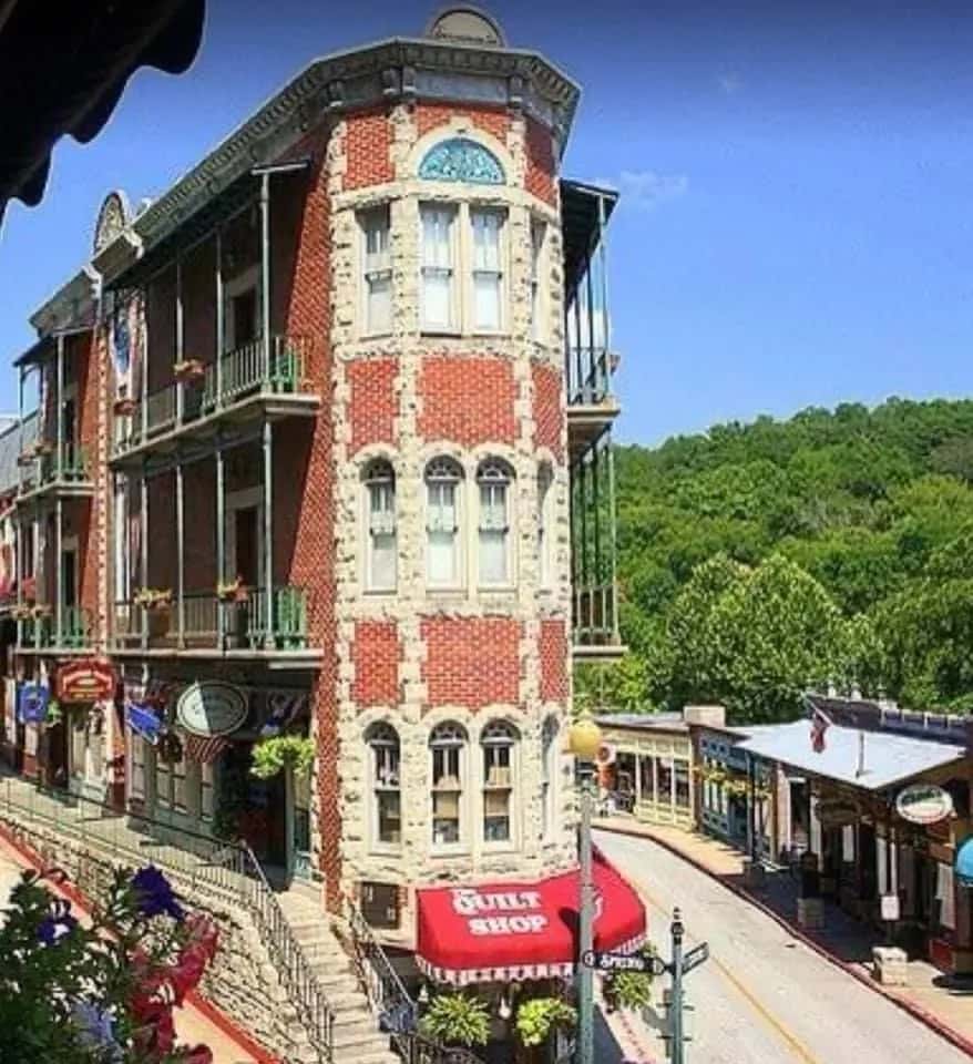 Historic red-brick flatiron-style building with ornate windows and balconies, housing shops on a quaint, sloped street lined with greenery.
