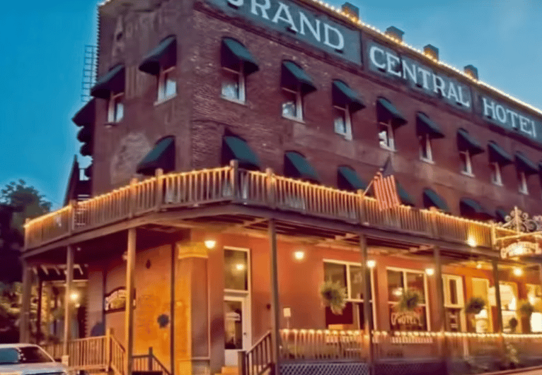 Brick exterior of the Grand Central Hotel lit by warm string lights, with green awnings and a wraparound porch at dusk.