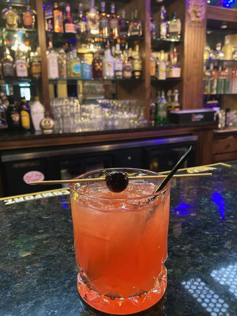 A vibrant reddish-orange cocktail served in a textured glass on the rocks, garnished with a dark cherry skewered on a wooden stick. The drink is placed on a glossy bar top, with a fully stocked bar visible in the background.
