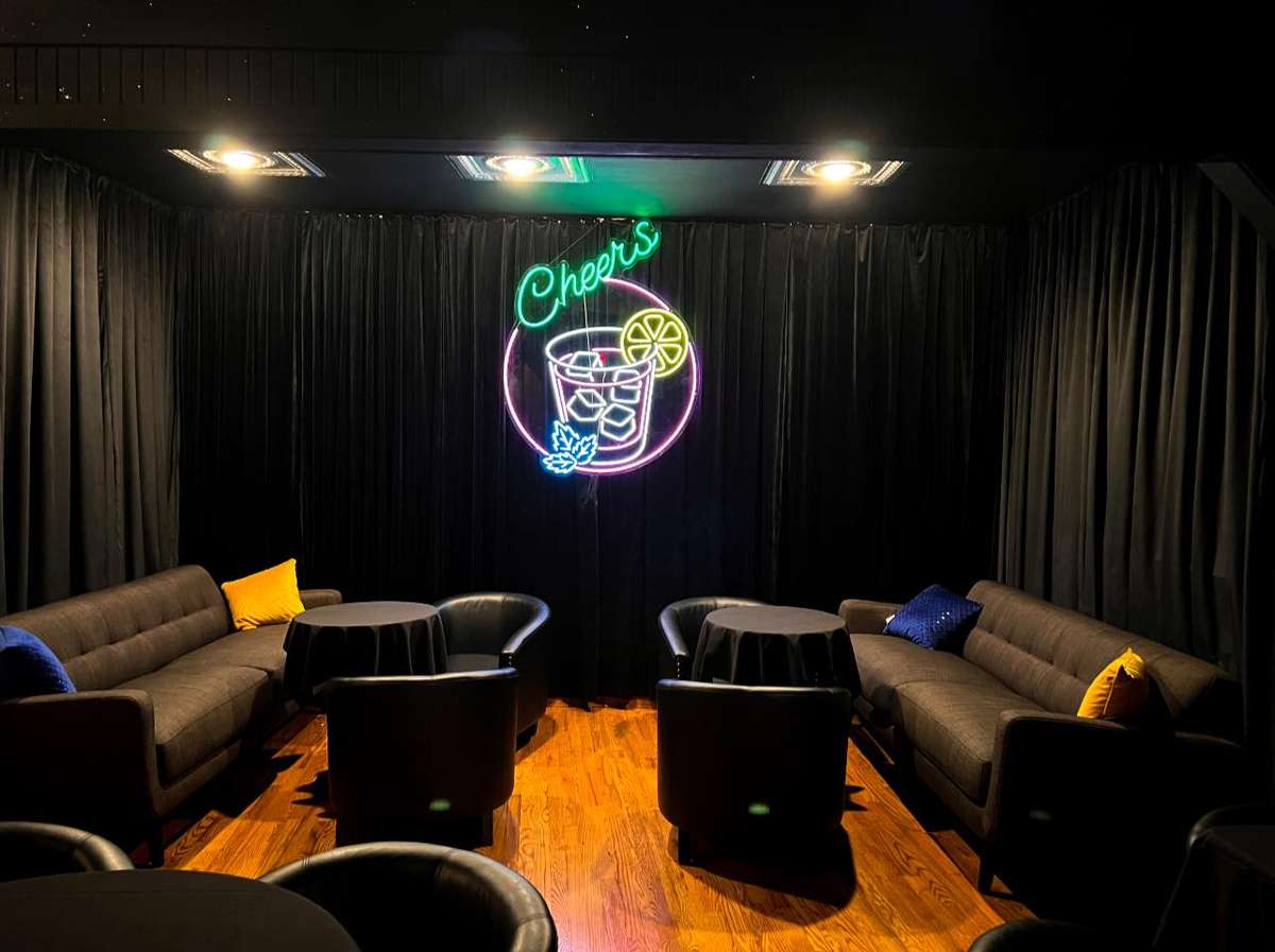 Cozy lounge area with black curtains, round tables, sofas, and a neon "Cheers" sign featuring a cocktail glass and lemon slice.