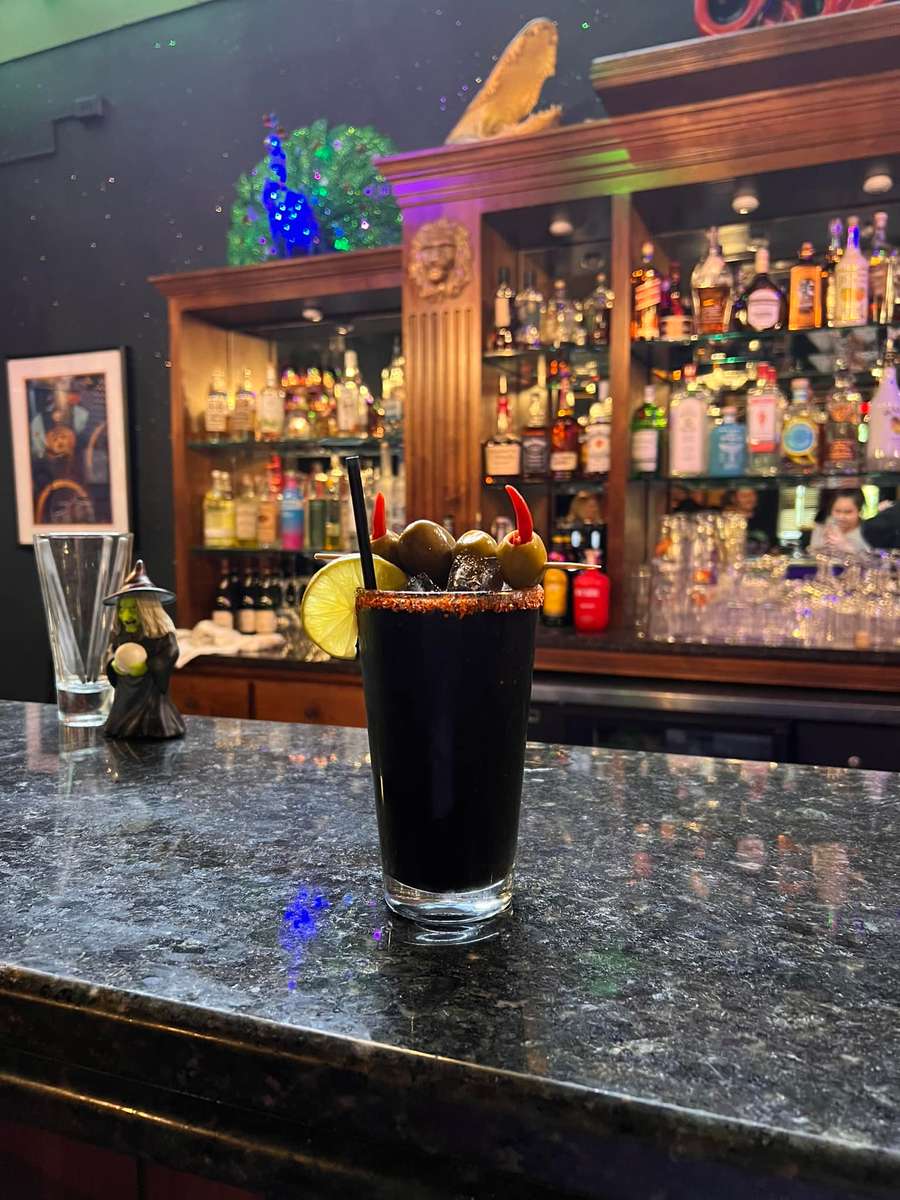 Dark cocktail garnished with olives, peppers, and lime on a bar counter, with a salt or spice-rimmed glass.
