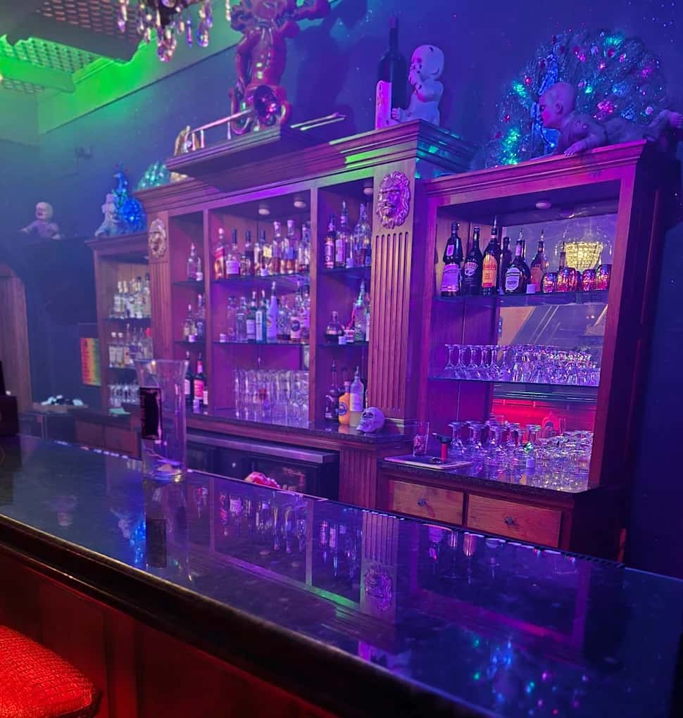 Eclectic bar setup with colorful lighting, vintage wooden shelves stocked with liquor, and quirky doll decorations throughout.
