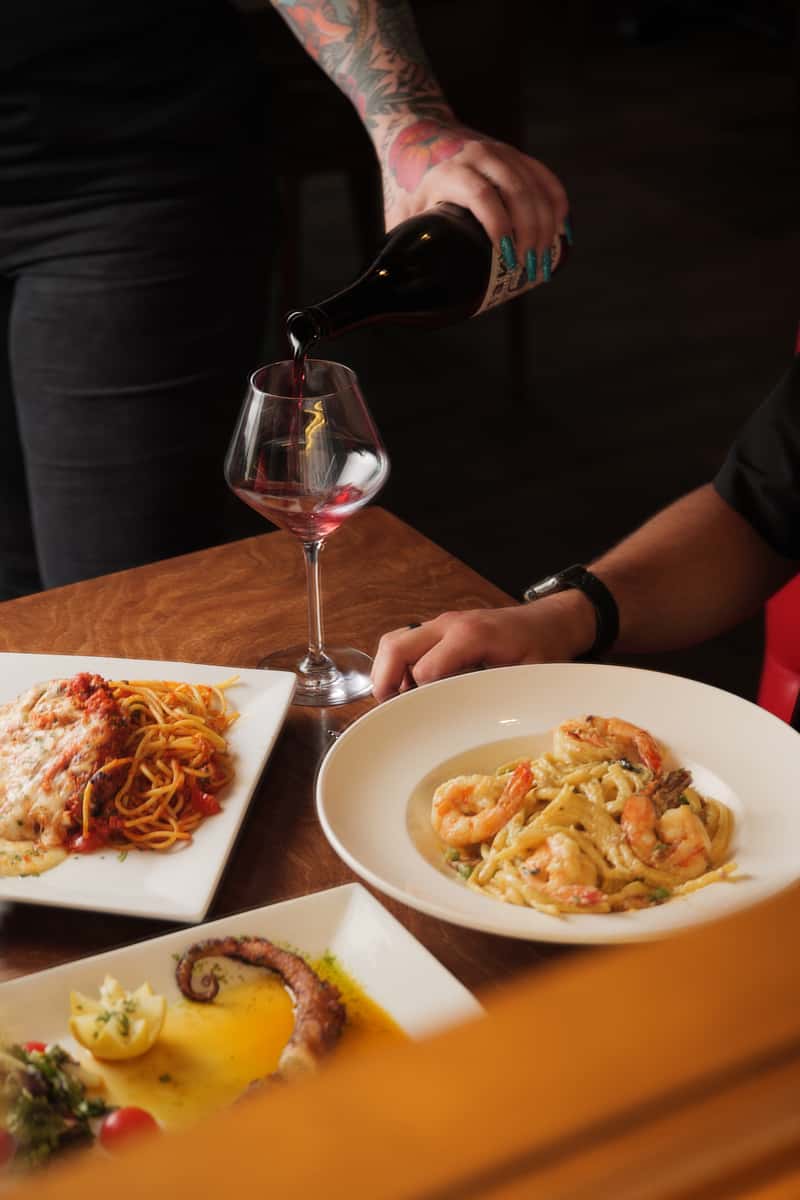 Server pouring red wine into a glass at an Italian restaurant table with shrimp pasta in creamy sauce, spaghetti marinara, and grilled octopus, capturing an upscale Italian dining experience.