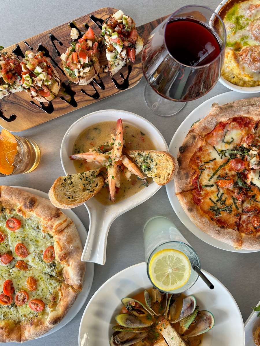 Overhead Italian restaurant spread with bruschetta, shrimp scampi, two wood-fired pizzas, baked meatballs, cocktails, and red wine.