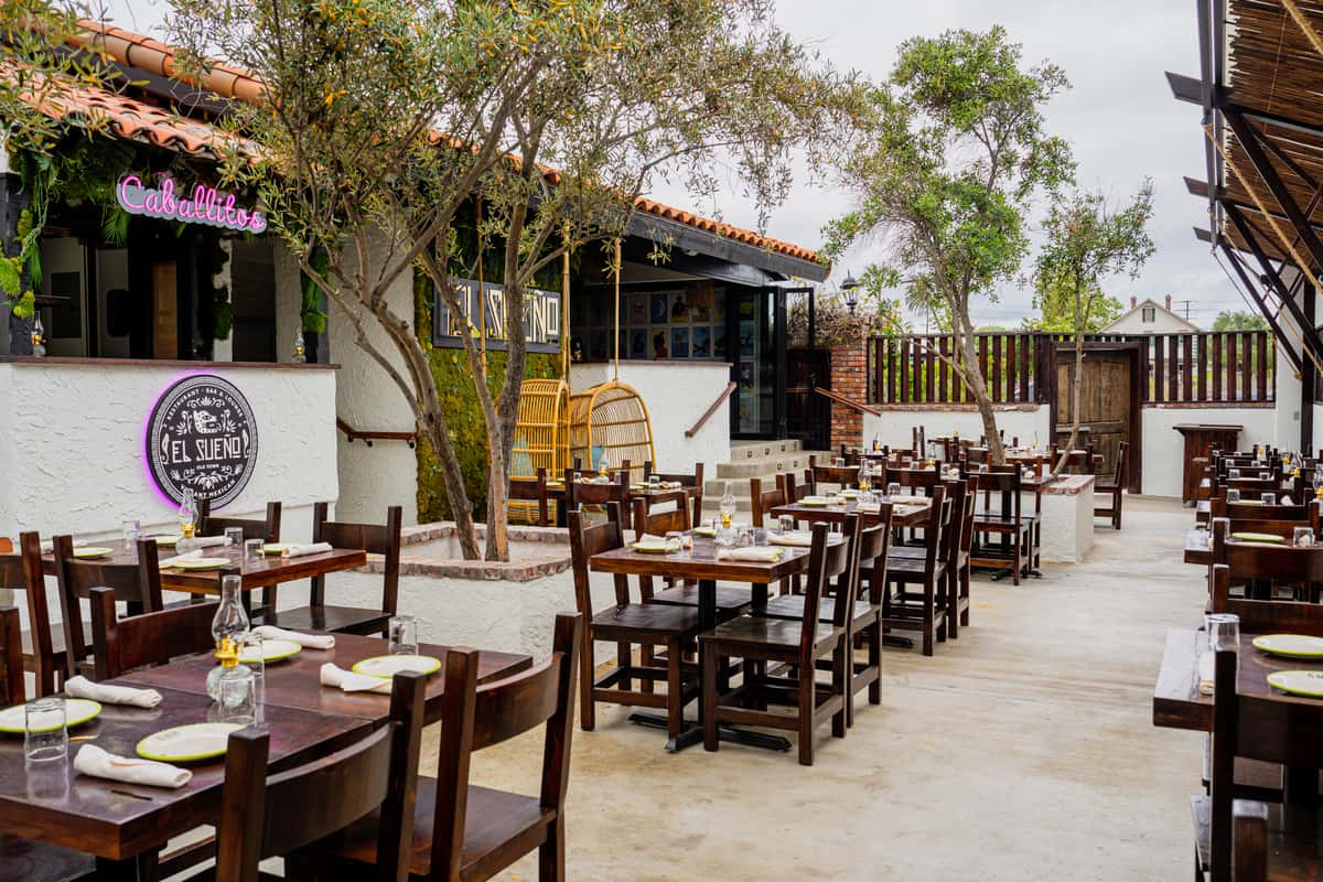 Al fresco charm at El Sueno: Outdoor patio with wooden tables, matching chairs, white stucco walls, and lush greenery.
