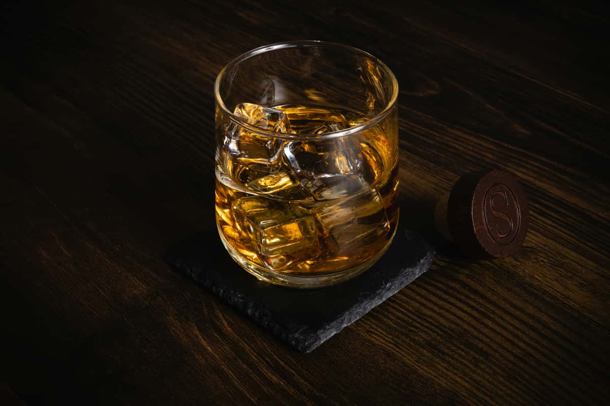 A glass of whiskey served over large ice cubes, resting on a black slate coaster on a dark wooden table, evoking a refined cocktail lounge ambiance.