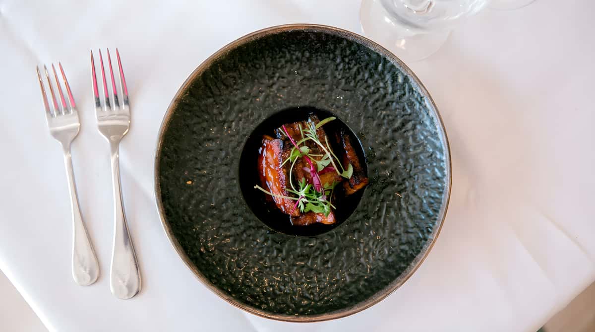 Fine dining plate with pork belly and microgreens in a black bowl.