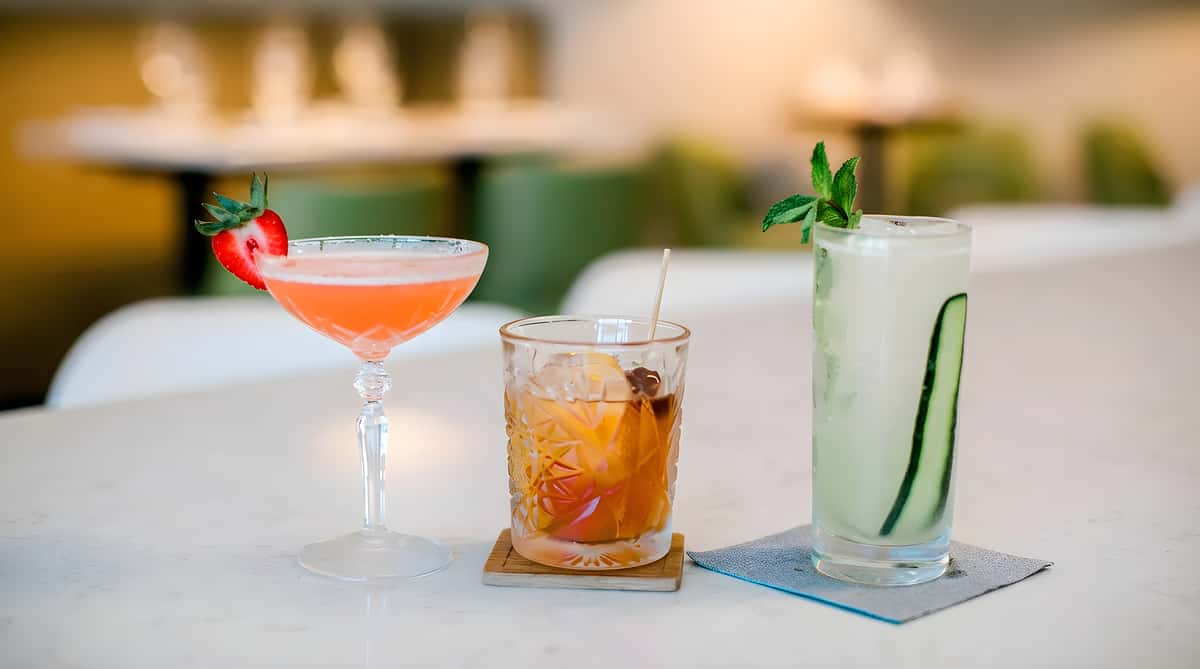 Three cocktails on a bar table, including a martini, old fashioned, and tall green drink.