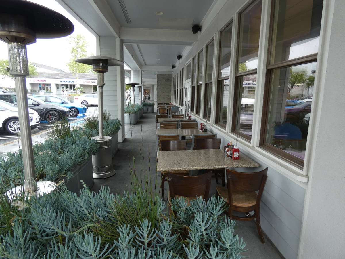 Covered outdoor dining patio with granite-top tables, wooden chairs, heat lamps, and lush succulent planters facing a parking lot.