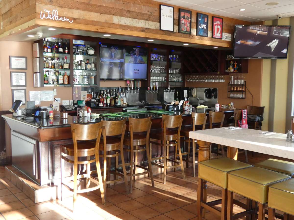 Sunlit sports bar with a polished wood counter, high chairs, liquor shelves, flat-screen TVs, and framed jerseys on a rustic wall.