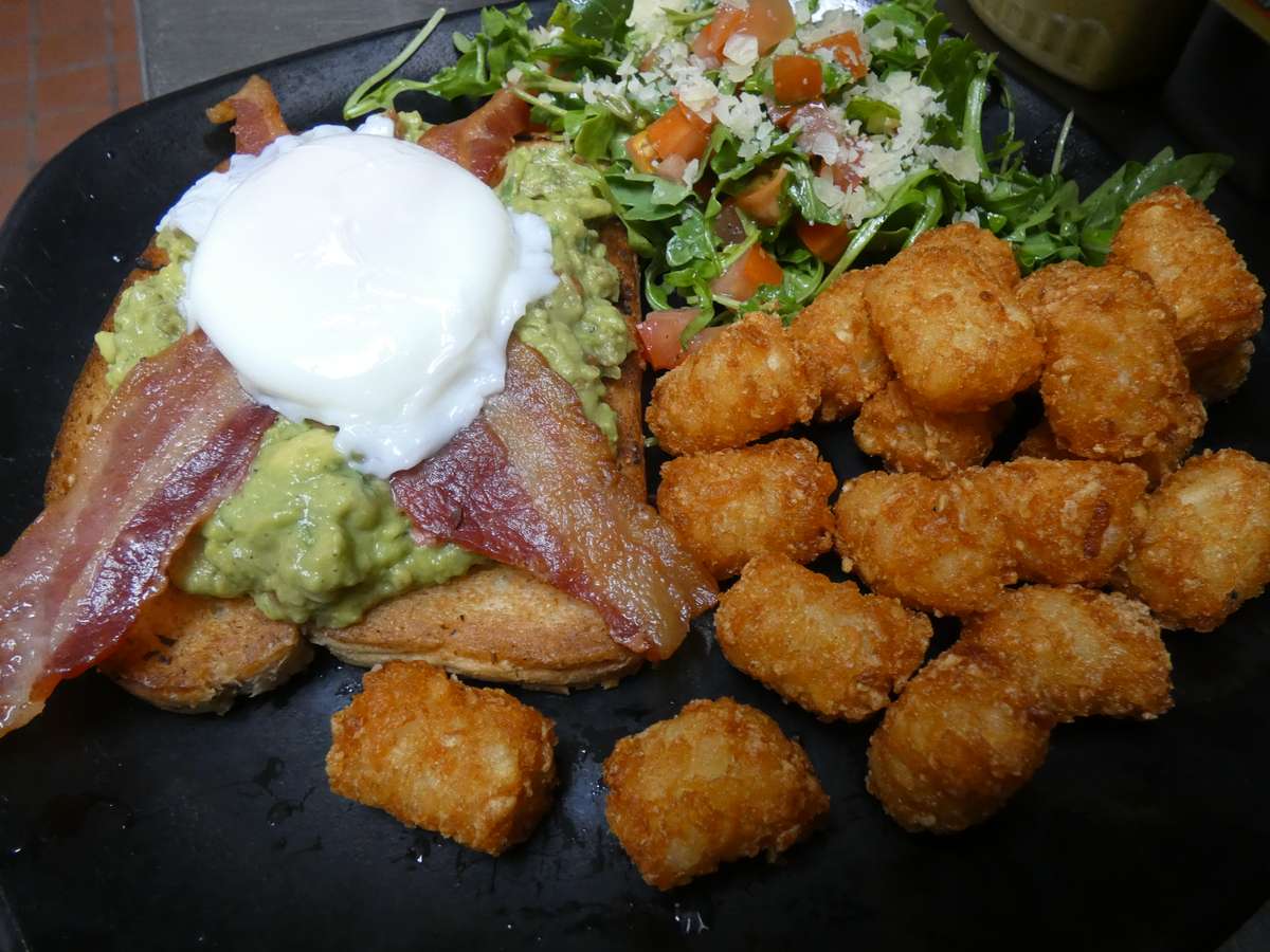 Brunch plate with avocado toast topped with bacon and a poached egg, crispy tater tots, and arugula salad with diced tomatoes.