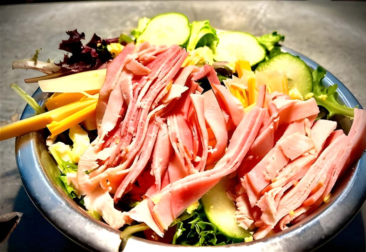 A large bowl filled with mixed greens, sliced deli meats (ham and turkey), cucumbers, and shredded cheese.