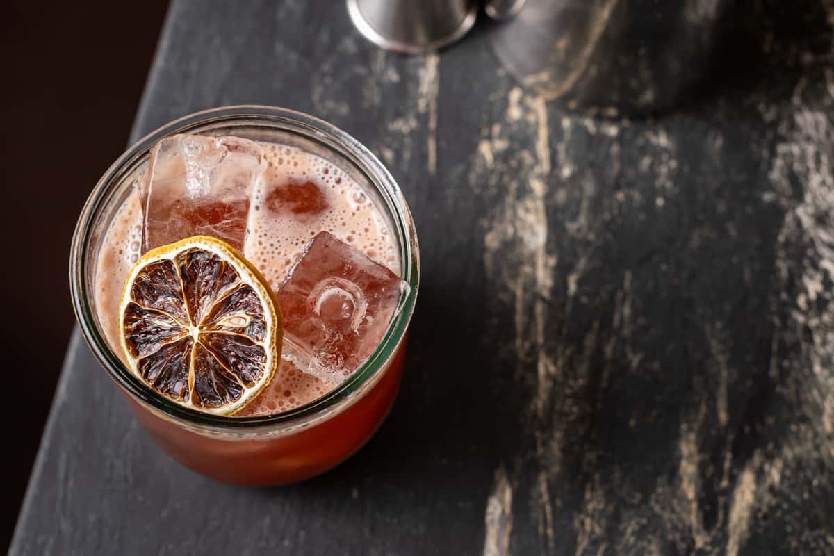 Top-down view of a mezcal cocktail with a smoky reddish hue, served over large ice cubes and garnished with a dehydrated citrus wheel in a short glass on a dark stone bar.