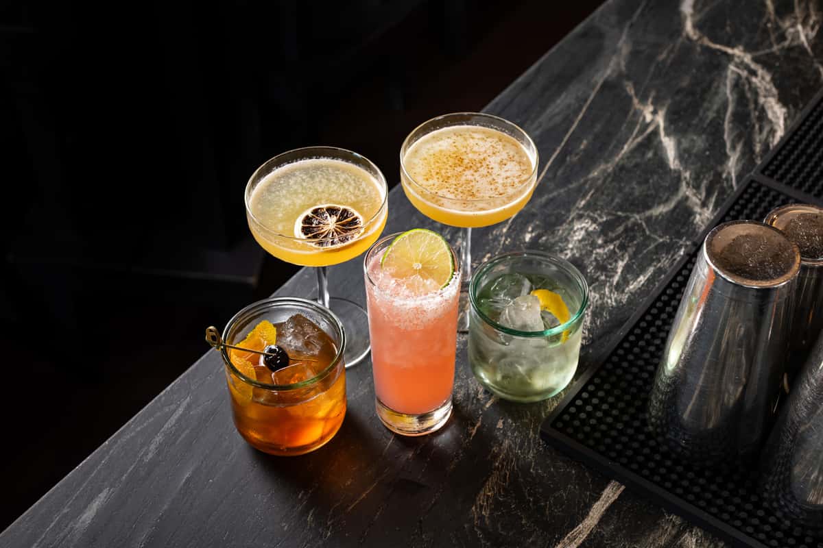 Curated lineup of five craft cocktails on a dark marble bar at La Holly, featuring mezcal and tequila-based drinks in coupe, highball, and rocks glasses with citrus and cherry garnishes