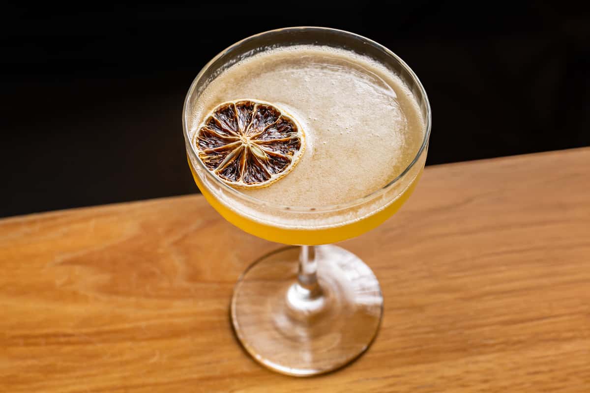 Elegant citrus cocktail in a coupe glass, garnished with a dehydrated lime wheel, served on a polished wooden bar top.