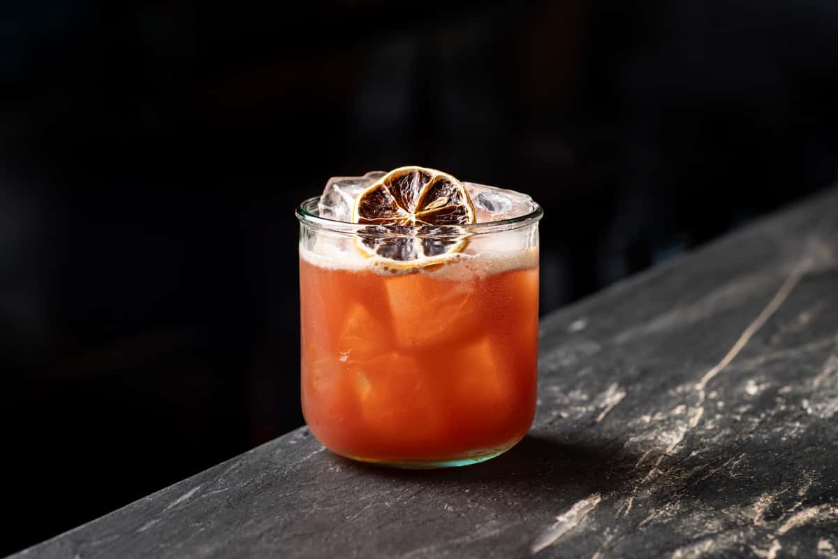 Craft cocktail in a short glass with a burnt citrus garnish, featuring a vibrant reddish-orange hue and large ice cubes on a dark marble bar
