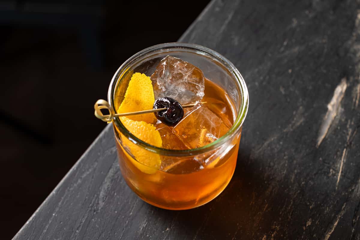 Classic Old Fashioned cocktail in a short glass with a large ice cube, garnished with orange peel and a cocktail cherry on a skewer, served on a dark stone bar.
