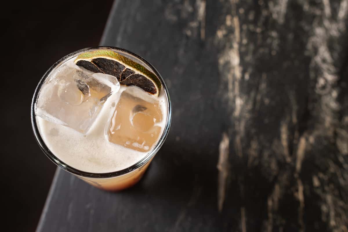 Overhead view of a refreshing mezcal cocktail in a tall glass with large ice cubes, frothy top, and a charred lime garnish, served on a rustic dark stone bar.