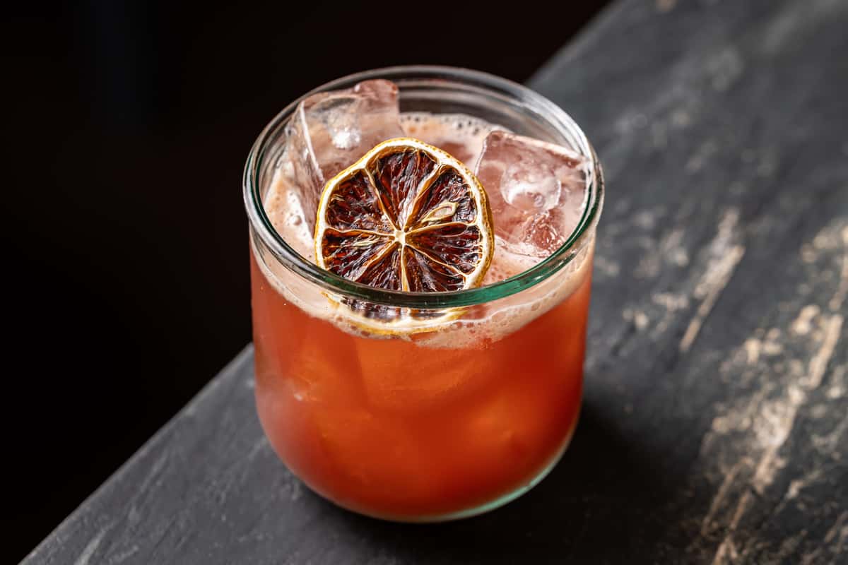 Vibrant mezcal cocktail with a smoky red hue, served in a short glass over ice and garnished with a charred citrus wheel, set on a dark stone bar at La Holly.
