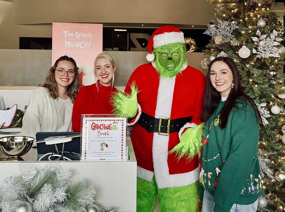 The Grinch stands next to Rutledge staff at the hostess stand for Grinchmas brunch event