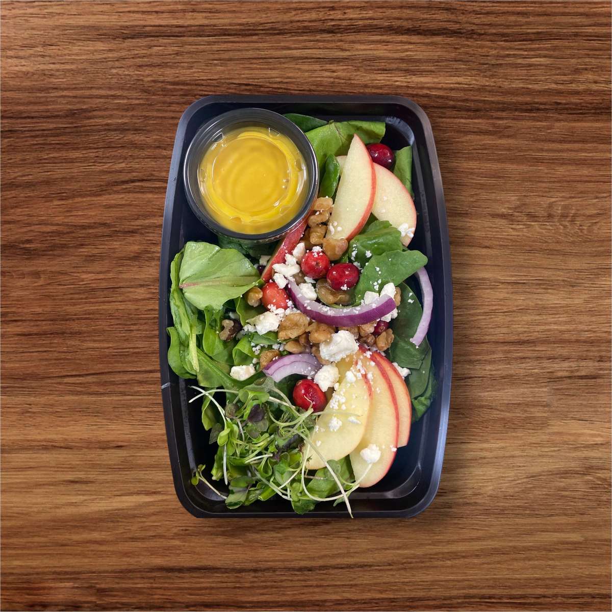 Fresh salad with spinach, apple slices, cranberries, red onion, feta, walnuts, and a cup of dressing in a black meal prep container.