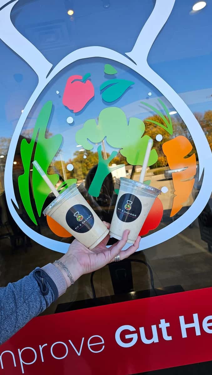 Two Fit & Savage smoothies held in front of a colorful gut health-themed window display with fruits and vegetables.