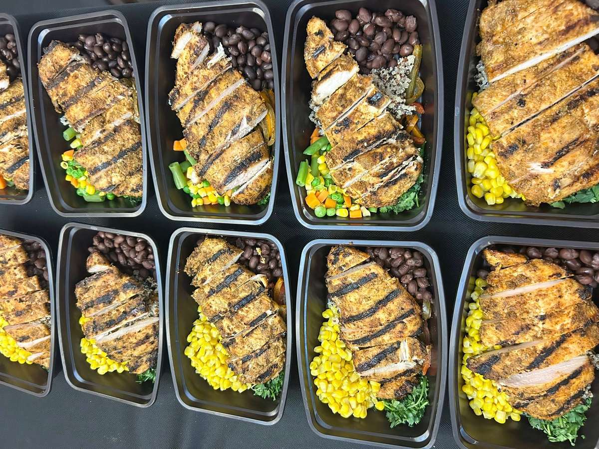 Grilled chicken breast meal prep with corn, black beans, and mixed vegetables in black containers.