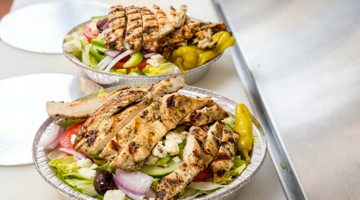 Two Greek salads with grilled chicken in foil containers, featuring lettuce, tomatoes, cucumbers, red onions, feta, black olives, and pepperoncini.